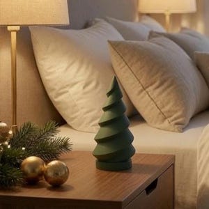 Könnte beinhalten: Eine matte, dunkelgrüne, spiralförmige Weihnachtsbaumfigur aus Holz auf einem flachen, runden Sockel. Der Baum steht auf einem Nachttisch aus Holz neben zwei goldenen, kugelförmigen Ornamenten und einem kleinen Zweig aus Kiefernnadeln.