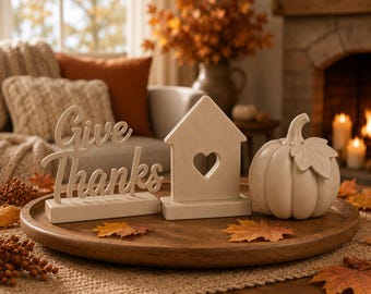May include: A wooden tray holds three decorative items: a beige sign with the cursive text "Give Thanks," a beige house-shaped ornament featuring a central heart cutout, and a beige ceramic pumpkin with a single leaf on its stem.