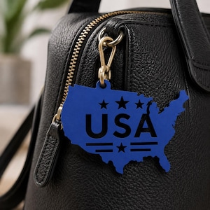 Puede incluir: Un colgante para bolso azul con la forma del mapa de los Estados Unidos, que presenta el texto USA en negro, tres estrellas sobre el texto y dos líneas horizontales con una estrella central debajo, sujeto a un bolso negro texturizado con cremallera dorada.