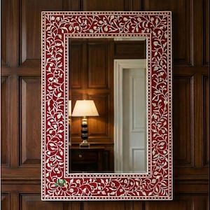 May include: A rectangular mirror with a red frame and white floral detailing. The mirror is mounted on a dark wood panelled wall. The frame has a detailed pattern of white leaves and dots, creating an elegant aesthetic.