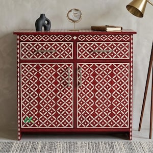 Może przedstawiać: Czerwona szafka z geometrycznym białym wzorem. Szafka posiada dwie szuflady i dwoje drzwi ze srebrnymi uchwytami. Na górze szafki znajdują się elementy dekoracyjne, w tym czarna rzeźba i złota lampa.