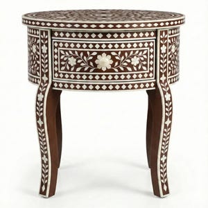 Puede incluir: Una mesa auxiliar redonda, marrón oscuro, con intrincados incrustaciones blancas de patrones florales y geométricos. La mesa presenta un solo cajón y patas curvas, mostrando una artesanía detallada. El diseño de la mesa es una mezcla de elegancia y arte.