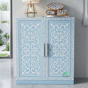 Puede incluir: Un armario azul claro con tallados florales blancos. El armario tiene dos puertas con tiradores plateados y un borde decorativo. La parte superior del armario tiene una bandeja plateada con objetos decorativos. El armario mide aproximadamente 76 cm de alto.