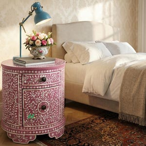 Könnte beinhalten: Ein runder, magentafarbener Nachttisch mit floralen Intarsien, mit einer Schublade und einer Schranktür. Eine blaue Schreibtischlampe und ein Blumenarrangement stehen darauf. Im Hintergrund ein Bett mit weißer Bettwäsche und einem beigen Kopfteil. Durchmesser ca. 50 cm.