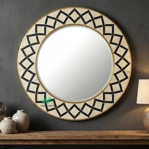 Op de afbeelding: Een ronde decoratieve spiegel met een goudkleurige lijst en een geometrisch zwart-wit patroon langs de rand. De spiegel hangt aan een donkergrijze muur boven een houten tafel. Een lamp en diverse decoratieve vazen staan op de tafel.