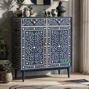 Puede incluir: Un mueble azul marino con dos cajones y dos puertas. El mueble está decorado con un estampado floral blanco. El mueble tiene pomos dorados y está sobre cuatro patas cónicas. El mueble está colocado frente a una pared con plantas.