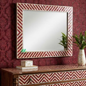 May include: A rectangular mirror with a red and white chevron patterned frame hangs above a matching chest of drawers. The chest has a similar chevron design. A vase with green plants sits on the chest.
