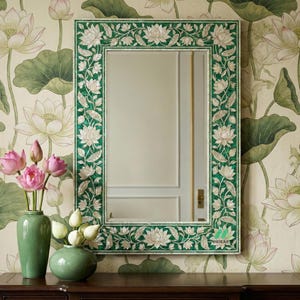 Puede incluir: Un espejo rectangular con un marco floral verde y blanco. El espejo se muestra sobre una superficie de madera oscura, con un jarrón de flores rosas y un jarrón de flores blancas. El fondo presenta un papel pintado floral.