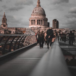 May include: A grayscale image of St. Paul's Cathedral in London, England, with people walking on a bridge in the foreground. The cathedral features a large dome and intricate architectural details. The sky is overcast.