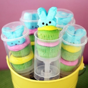 May include: A set of six clear plastic tubes filled with colorful cupcakes topped with blue and yellow marshmallow peeps. The cupcakes are decorated with green, yellow, and pink frosting. The tubes are arranged in a yellow bucket.