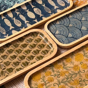 May include: Four rectangular wooden trays with decorative patterns. The trays feature designs including fish, wave-like patterns, and floral motifs in shades of blue, green, and yellow. The trays have a natural wood border.