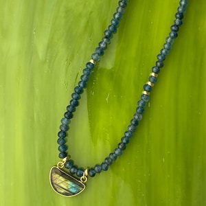 May include: A necklace featuring small, faceted, dark blue beads interspersed with tiny gold-toned spacers. The piece includes a gold-toned, semi-circular pendant with a dark, iridescent stone inlay that displays flashes of yellow and blue light.