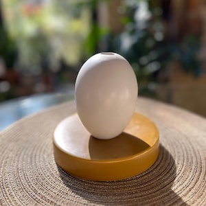 May include: A white, egg-shaped ceramic vase sits atop a round, mustard-colored base. The vase has a small opening at the top. The base is a solid color, and the vase is smooth. The items are on a textured, neutral-toned surface.