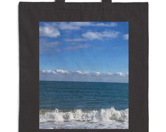 Puede incluir: Bolso tote negro con una imagen cuadrada de un paisaje marino. La imagen muestra un océano azul con olas blancas y un cielo azul con nubes blancas. El bolso tote tiene dos asas negras.