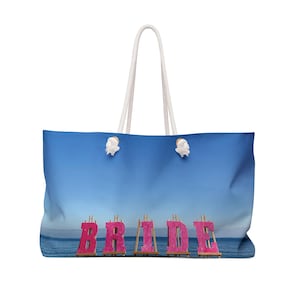May include: Large tote bag with a blue gradient background and white rope handles. The word "BRIDE" is displayed in pink letters. The bag is designed for carrying items and has a beach theme.