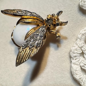 May include: A vintage bee brooch with gold-tone metal wings, accented with clear rhinestones and a large, iridescent white pearl body. The brooch has intricate detailing and a classic design, suitable for adding a touch of elegance to any outfit.
