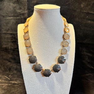 May include: A gold-toned necklace with alternating round, faceted beads. The beads transition from light peach to gray, creating a gradient effect. The necklace is displayed on a white mannequin.