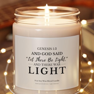 May include: A lit candle in a clear glass jar with a white label. The label reads "GENESIS 1:3 AND GOD SAID 'Let There Be Light.' AND THERE WAS LIGHT." The candle sits on a small wooden coaster, with string lights and a guitar in the background.