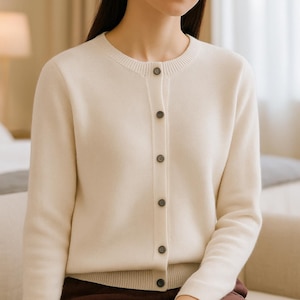 May include: A cream-coloured cardigan with a round neckline and button-down front. The long-sleeved sweater is paired with dark burgundy trousers. The buttons are a dark brown colour. The model is seated on a light-coloured sofa.