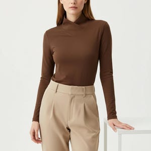 May include: A long-sleeved brown top with a unique neckline and a pair of beige pants. The top has a high, asymmetrical collar. The pants are high-waisted with a pleated front. The outfit is simple and elegant.