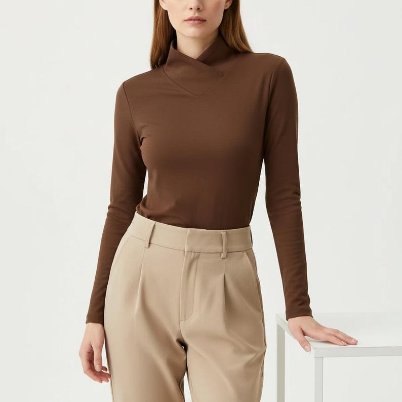 May include: A long-sleeved brown top with a unique neckline and a pair of beige pants. The top has a high, asymmetrical collar. The pants are high-waisted with a pleated front. The outfit is simple and elegant.