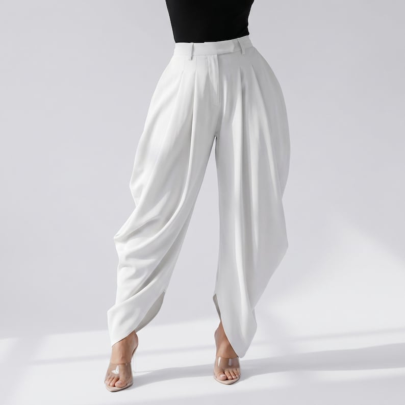 May include: White high-waisted trousers with a distinctive draped design. The trousers feature a wide leg and a tailored waistband. The model is wearing clear heeled sandals and a black top. The trousers are made of a smooth, flowing fabric.