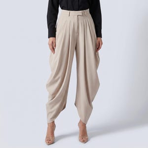 May include: Beige high-waisted trousers with a distinctive draped design. The trousers feature a tailored waistband and pleats, creating a voluminous silhouette. The model is wearing a black long-sleeved shirt and clear heels. The trousers are made of a smooth, flowing fabric.