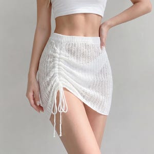 May include: A white mini skirt crafted from a semi-sheer knit fabric. The skirt features a high-waisted design with a ribbed waistband and a vertical drawstring detail on the left side that creates a gathered, ruched effect with hanging ties.