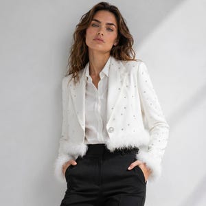 Pode incluir: Um blazer branco curto com strass prateados espalhados, fecho de botão de strass único e acabamento em penas brancas na bainha e nos punhos, usado sobre uma camisa branca de colarinho e calças pretas.