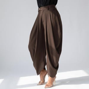 May include: Brown, high-waisted trousers with a distinctive, voluminous design. The trousers feature pleats at the waist and a wide, draped leg. The model is wearing clear high heels. The trousers are made of a smooth, flowing fabric.