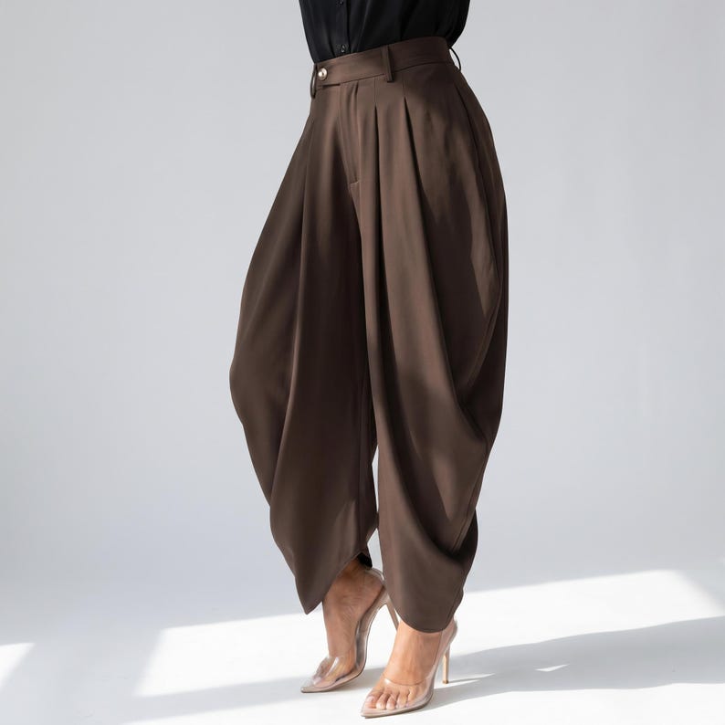 May include: Brown, high-waisted trousers with a distinctive, voluminous design. The trousers feature pleats at the waist and a wide, draped leg. The model is wearing clear high heels. The trousers are made of a smooth, flowing fabric.
