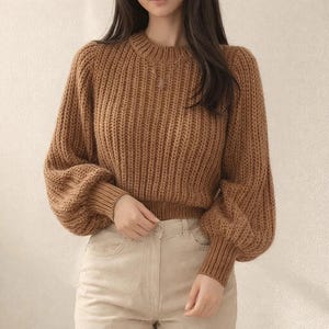 May include: A brown, chunky knit jumper with a crew neck and long, voluminous sleeves. The jumper has ribbed cuffs and a ribbed hem. The model is wearing the jumper with light-coloured trousers.