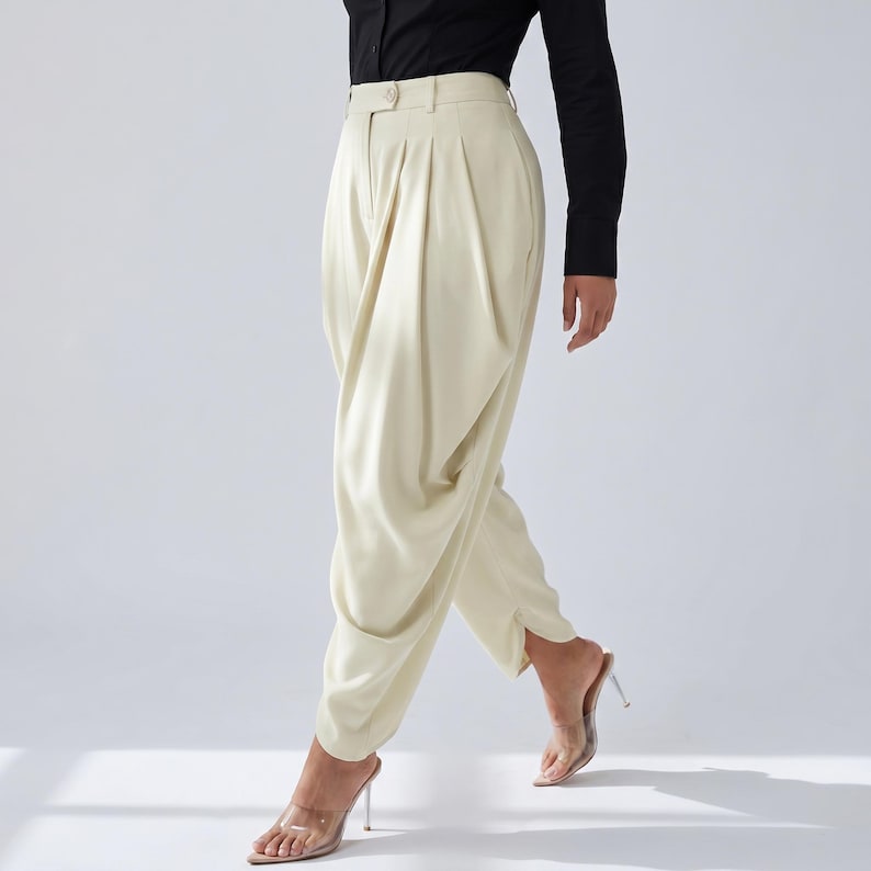 May include: Cream-coloured, high-waisted trousers with a distinctive draped design. The trousers have a button closure and pleats at the waist. The model is wearing a black long-sleeved shirt and clear heeled sandals.