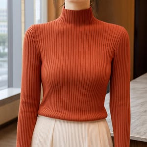 Può includere: Un maglione a collo alto a coste color arancione bruciato con maniche lunghe. Il maglione ha una vestibilità aderente e un collo alto. La parte inferiore dell'immagine mostra una gonna color crema. Il maglione è realizzato in un materiale morbido e confortevole.