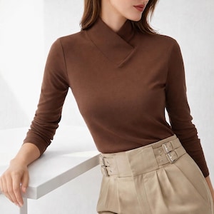 May include: A long-sleeved brown top with a unique neckline. The top is paired with beige pants featuring a wide belt with double silver buckles. The outfit is simple and elegant.
