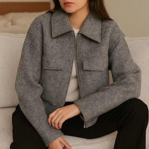 Könnte beinhalten: Graue Wolljacke mit Kragen und silbernem Reißverschluss. Die Jacke hat zwei Vordertaschen und wird über einem weißen Hemd getragen. Die Person trägt eine schwarze Hose.
