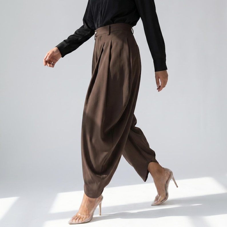 May include: Brown high-waisted trousers with pleats, worn with a black long-sleeved shirt. The trousers have a wide leg design. The model is wearing clear high heels. The outfit is set against a white background.