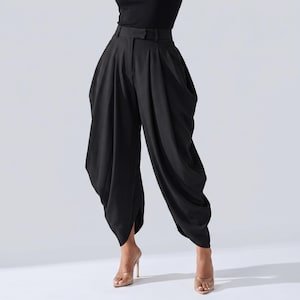 May include: Black high-waisted trousers with a distinctive, draped style. The trousers have a wide leg and a tailored waistband with a belt loop. The fabric looks to be a smooth, flowing material. The trousers are worn with clear high-heeled sandals.