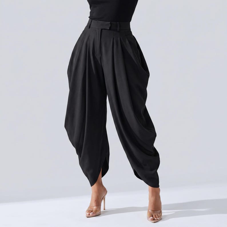 May include: Black high-waisted trousers with a distinctive, draped style. The trousers have a wide leg and a tailored waistband with a belt loop. The fabric looks to be a smooth, flowing material. The trousers are worn with clear high-heeled sandals.