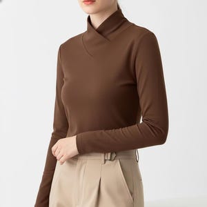 May include: A long-sleeved brown top with a unique neckline. The top has a high collar that folds over into a V-neck. The sleeves are long and fitted. The model is wearing tan pants with a belt.