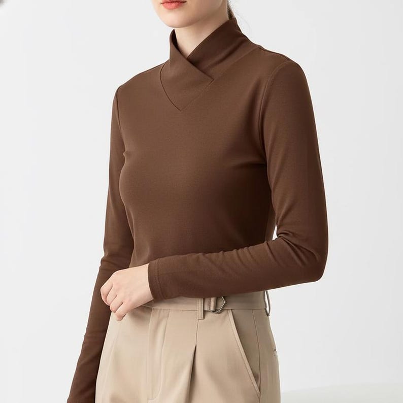 May include: A long-sleeved brown top with a unique neckline. The top has a high collar that folds over into a V-neck. The sleeves are long and fitted. The model is wearing tan pants with a belt.