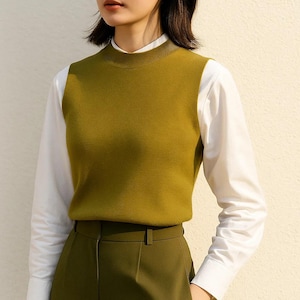 May include: A woman wearing a mustard-colored knit vest over a white collared shirt. The vest has a mock neck and is paired with olive green trousers. The outfit is simple and stylish, suitable for a casual or professional setting.