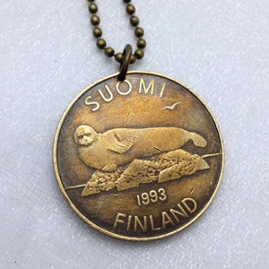 May include: A bronze necklace with a round pendant featuring a seal on a rock with the text "SUOMI 1993 FINLAND".