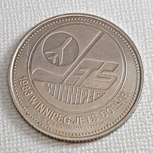以下が含まれることがあります： 1983年の銀貨、ウィニペグ・ジェッツ・ダラー。コインには、ウィニペグ・ジェッツのロゴと、様式化されたジェット機と「ウィニペグ・ジェッツ・ダラー」の文字が描かれています。コインの縁にはテクスチャパターンがあります。