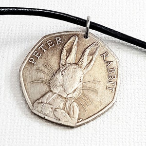 May include: A silver pendant featuring Peter Rabbit from the children's book series. The pendant is a coin with the words "PETER RABBIT" around the edge and a detailed image of the rabbit. It hangs from a black cord.