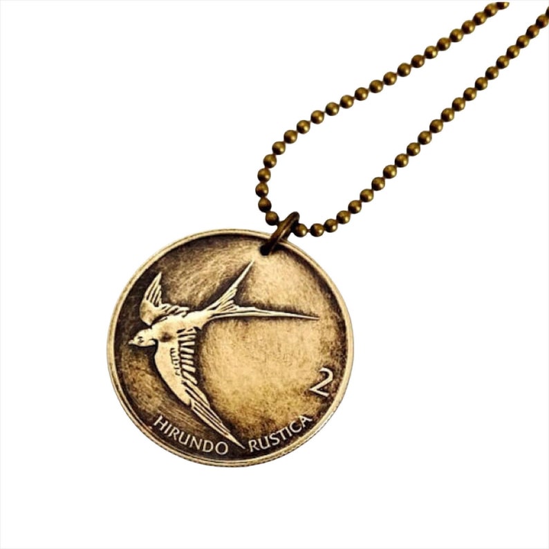 May include: A gold-toned necklace with a round pendant featuring a stylized image of a swallow in flight. The pendant is inscribed with the words "Hirundo Rustica" and the number "2".