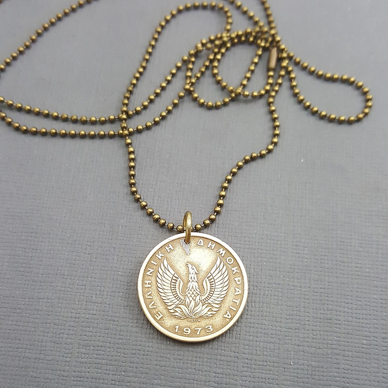 May include: A gold chain necklace with a pendant featuring a Greek coin from 1973. The coin has a phoenix design and the text "&Epsilon;&Lambda;&Lambda;&Eta;&Nu;&Iota;&Kappa;&Eta; &Delta;&Eta;&Mu;&Omicron;&Kappa;&Rho;&Alpha;&Tau;&Iota;&Alpha;".