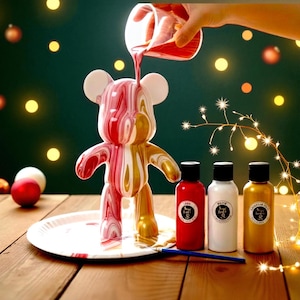 May include: A decorative bear figurine with red, white, and gold paint being poured onto it. The bear is on a white plate, with three paint bottles and a paintbrush nearby. The background is a dark green with Christmas lights.