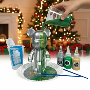 May include: A silver and green bear figurine being painted with green paint. The figurine is on a white plate with a paintbrush. Paint bottles and a mixing cup are also present. The background includes a Christmas tree and a fireplace.