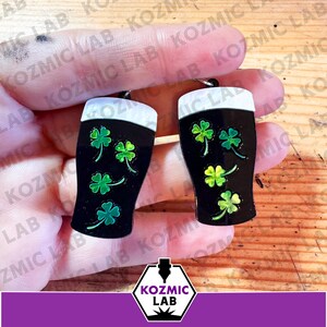 May include: A pair of black beer glass earrings with white foam tops and green shamrock designs. The earrings are held up against a wooden background. The earrings are a St. Patrick's Day theme.